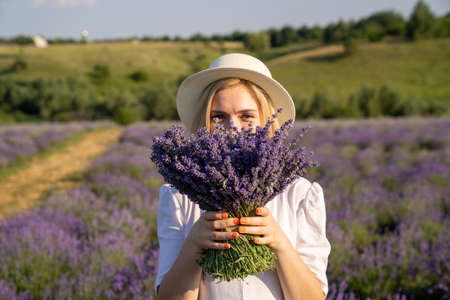 portrait of woman model in white dress outfit with hat is standing in lavender field, photo session. Young girl is holding lavender bouquet. Good picture for cover, calendarの写真素材