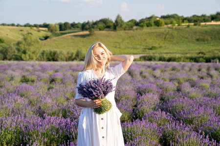 woman model in white dress outfit is standing in lavender field with purple bouquet, photo session. Young girl is holding lavender bouquet. Portrait of  girl touching her hairの写真素材