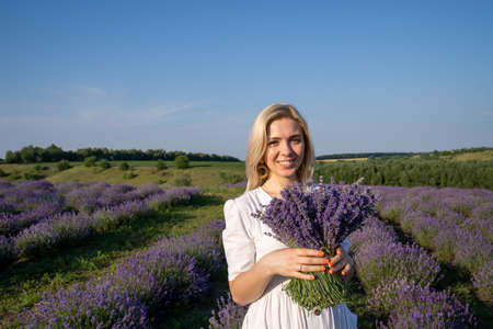 portrait of woman model in white dress outfit with hat is standing in lavender field, photo session. Young girl is holding lavender bouquet. Good picture for cover, calendarの写真素材