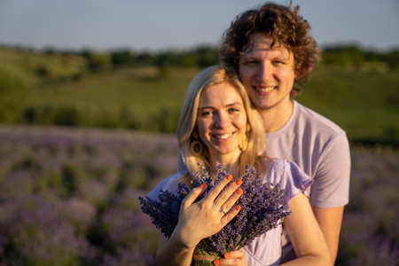 portrait of a couple man and woman in matching lilac outfit. Girl is holding lavender bouquet and there is diamond engagement ring on her hand. They are happy and hugの写真素材