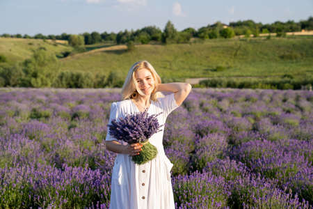 portrait of woman model in white dress outfit with hat is standing in lavender field, photo session. Young girl is holding lavender bouquet. Good picture for cover, calendarの写真素材