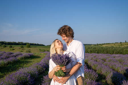 portrait of a couple in matching white outfit. Girl is holding lavender bouquet and smiling. They are happy and hug showing emotions. Good picture for cover, calendar, postcardの写真素材