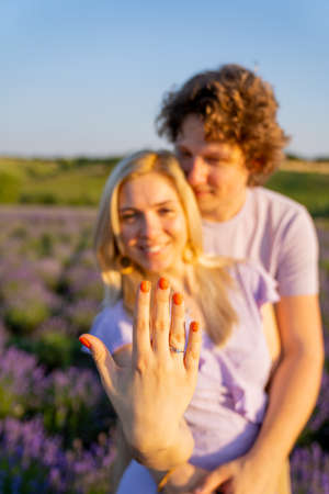 portrait of a couple man and woman in matching lilac outfit. Girl is holding lavender bouquet and there is diamond engagement ring on her hand. They are happy and hugの写真素材
