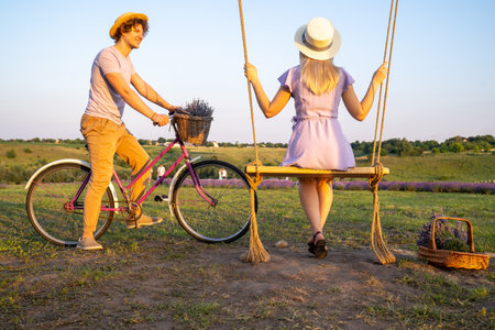 young couple in love, woman is sitting on the swing, man is riding bicycle on the lavender field, sunset. Concept of romantic relationship and dateの写真素材