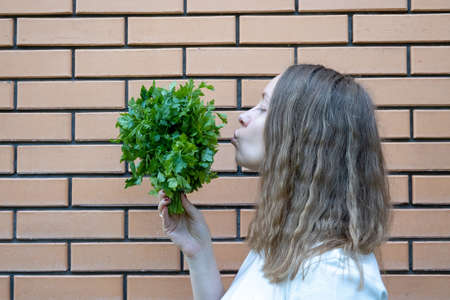 young woman in white t shirt with green parsley bunch in her hands. She holds parsley in front of her face, making fun, standing next to brick wall. Positive emotions, She is tryinの写真素材
