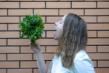 young woman in white t shirt with green parsley bunch in her hands. She holds parsley in front of her face, making fun, standing next to brick wall. Positive emotions, She is tryinの写真素材