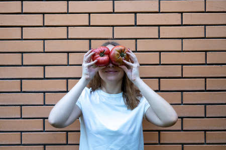 young woman in white t shirt with red tomatoes in her hands. She holds tomatoes in front of her eyes, making fun face, standing next to brick wall. Positive emotionsの写真素材