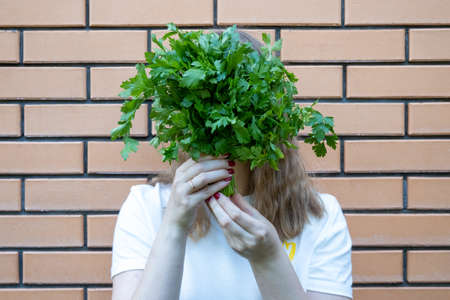 young woman in white t shirt with green parsley bunch in her hands. She holds parsley in front of her face, making fun, standing next to brick wall. Positive emotions, She is tryinの写真素材