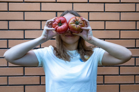 young woman in white t shirt with red tomatoes in her hands. She holds tomatoes in front of her eyes, making fun face, standing next to brick wall. Positive emotionsの写真素材