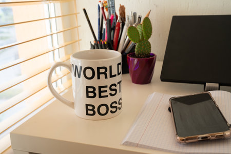 blur effect, workplace table with cup "world's best boss" of coffee, closeup of office supplies, stationary, plant orchid, cactus, smartphone near laptop, computer mouseの写真素材