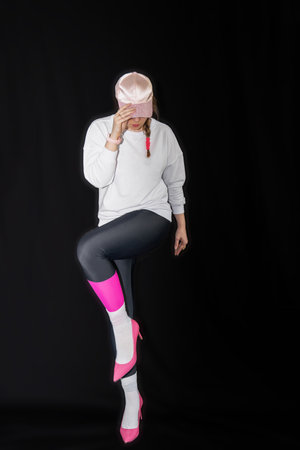 young woman wearing trendy clothes isolated on a black background. Stylish model in white sweatshirt, black leggings, white socks and pink heels shoes, with pink cap is posingの写真素材
