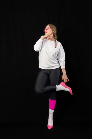 young woman wearing trendy clothes isolated on a black background. Stylish model in white sweatshirt, black leggings, white socks and pink heels shoes, with pink glasses is posingの写真素材