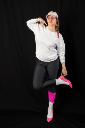 young woman wearing trendy clothes isolated on a black background. Stylish model in white sweatshirt, black leggings, white socks and pink heels shoes, with pink cap is posingの写真素材