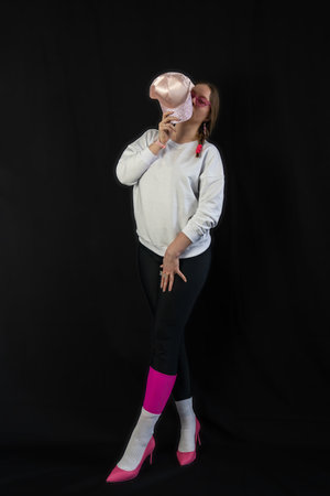 young woman wearing trendy clothes isolated on a black background. Stylish model in white sweatshirt, black leggings, white socks and pink heels shoes, with pink cap is posingの写真素材