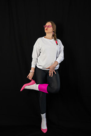 young woman wearing trendy clothes isolated on a black background. Stylish model in white sweatshirt, black leggings, white socks and pink heels shoes, with pink glasses is posingの写真素材