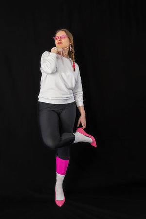 young woman wearing trendy clothes isolated on a black background. Stylish model in white sweatshirt, black leggings, white socks and pink heels shoes, with pink glasses is posingの写真素材