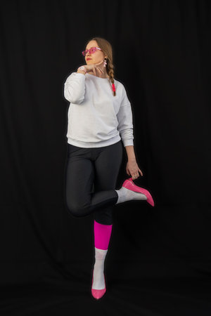 young woman wearing trendy clothes isolated on a black background. Stylish model in white sweatshirt, black leggings, white socks and pink heels shoes, with pink glasses is posingの写真素材