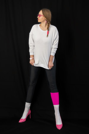young woman wearing trendy clothes isolated on a black background. Stylish model in white sweatshirt, black leggings, white socks and pink heels shoes, with pink glasses is posingの写真素材