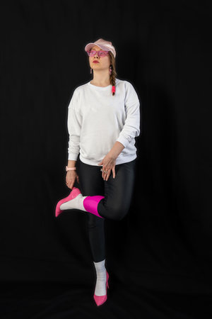young woman wearing trendy clothes isolated on a black background. Stylish model in white sweatshirt, black leggings, white socks and pink heels shoes, with pink cap is posingの写真素材