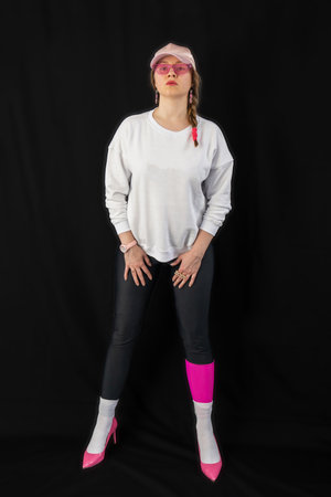 young woman wearing trendy clothes isolated on a black background. Stylish model in white sweatshirt, black leggings, white socks and pink heels shoes, with pink cap is posingの写真素材
