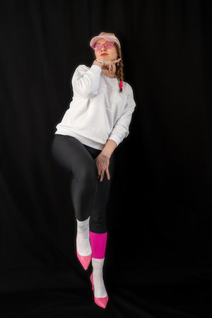 young woman wearing trendy clothes isolated on a black background. Stylish model in white sweatshirt, black leggings, white socks and pink heels shoes, with pink cap is posingの写真素材
