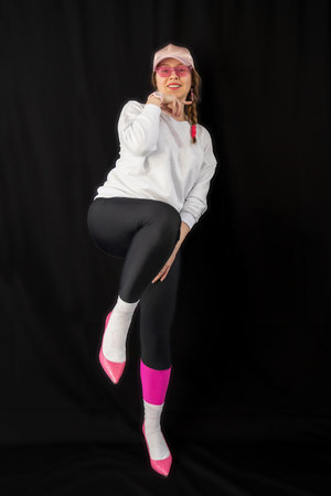 young woman wearing trendy clothes isolated on a black background. Stylish model in white sweatshirt, black leggings, white socks and pink heels shoes, with pink cap is posingの写真素材