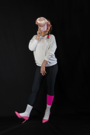 young woman wearing trendy clothes isolated on a black background. Stylish model in white sweatshirt, black leggings, white socks and pink heels shoes, with pink cap is posingの写真素材