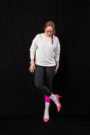 young woman wearing trendy clothes isolated on a black background. Stylish model in white sweatshirt, black leggings, white socks and pink heels shoes, with pink glasses is posingの写真素材