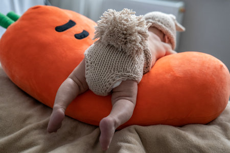 cute newborn baby in beige knitted rabbit costume is lying on orange plush pillow, infant is crawling on carrot toy, learning how to walkの写真素材