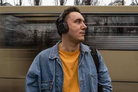 handsome black haired man in blue jeans jacket is standing on the bus stop, waiting for a bus. Person is listening to music in wireless headphones. Blurred motion of transportの写真素材