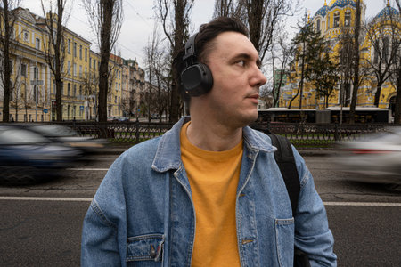 handsome black haired man in blue jeans jacket is standing on the street near heavy traffic. Person is listening to music in wireless headphones. Blurred motion of carsの写真素材