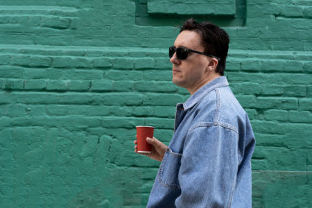 stylish man with dark black hair, wearing sunglasses and blue jeans jacket, standing outdoors next to green wall, holding plastic cup of drink. Street fashion, casual clothesの写真素材