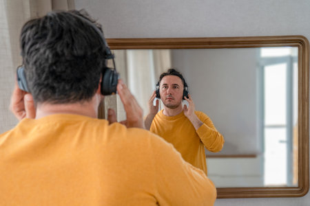 a handsome man with a beard in a yellow sweater outfit is looking in the mirror. Person wears headphones. Male is dressing for a walkの写真素材
