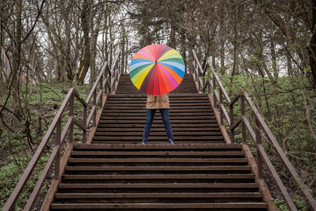 young woman in a beige coat and rain boots stands in forest or park and holds colorful bright rainbow umbrella. Autumn weather in city, girl is walking on a wooden stairsの写真素材