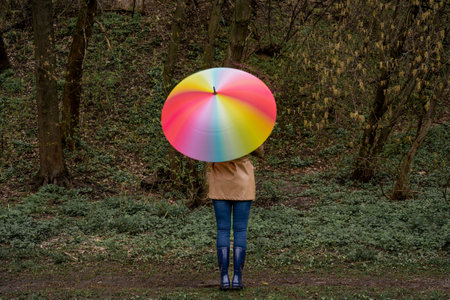 young woman in a beige coat and rain boots stands in forest or park and holds colorful bright rainbow umbrella. Girl spins umbrella for blur effect. Slow shutter speed. Autumnの写真素材