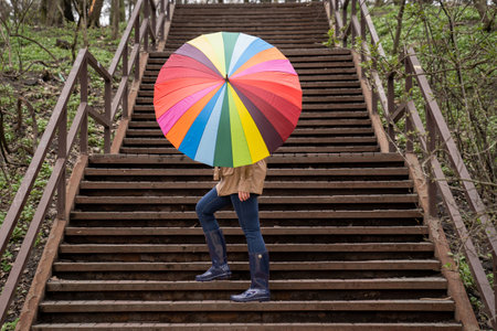 young woman in a beige coat and rain boots stands in forest or park and holds colorful bright rainbow umbrella. Autumn weather in city, girl is walking on a wooden stairsの写真素材