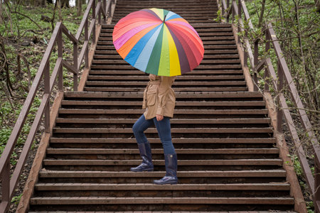 young woman in a beige coat and rain boots stands in forest or park and holds colorful bright rainbow umbrella. Autumn weather in city, girl is walking on a wooden stairsの写真素材