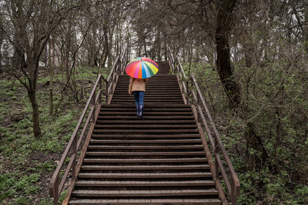 young woman in a beige coat and rain boots stands in forest or park and holds colorful bright rainbow umbrella. Autumn weather in city, girl is walking on a wooden stairsの写真素材