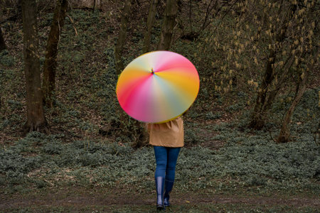 young woman in a beige coat and rain boots stands in forest or park and holds colorful bright rainbow umbrella. Girl spins umbrella for blur effect. Slow shutter speed. Autumnの写真素材