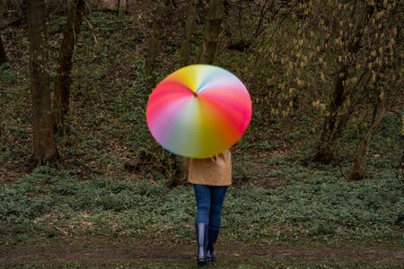 young woman in a beige coat and rain boots stands in forest or park and holds colorful bright rainbow umbrella. Girl spins umbrella for blur effect. Slow shutter speed. Autumnの写真素材