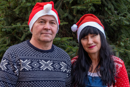 family in santa claus red hats standing near a green Christmas tree. Happy new year merry christmas. Middle aged in their 50th husband and wife wear festive sweater clothesの写真素材