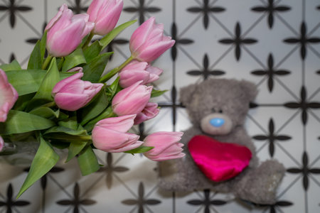 grey plush toy teddy bear with pink heart next to pink blooming flower bouquet of tulips in vase. Happy valentines day giftの写真素材