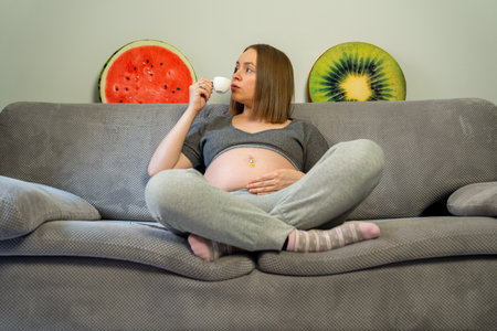 pregnant woman holds cup of hot drink and sits on sofa with plush pillows and drinks coffee. Caffeine during pregnancy. Expectant mother healthy lifestyle.の写真素材