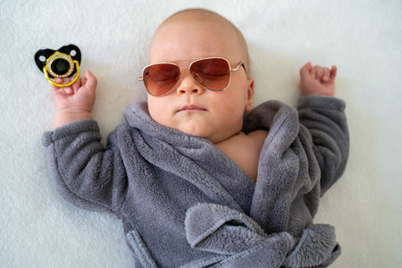 cute little newborn baby wearing sunglasses and grey bathrobe lying on a white background. Infant...