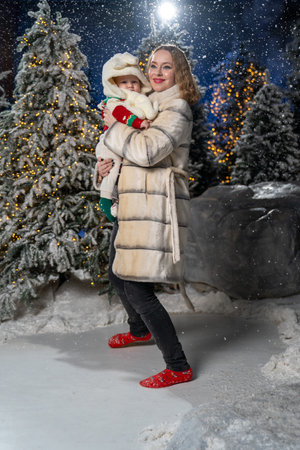 Full-body winter portrait of mother holding baby outdoors in snowy, festive setting. Mother is wearing a long beige puffer coat, black jeans, and red winter shoes, while baby is drの写真素材