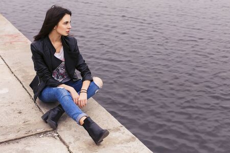 beautiful brunette woman in black leather jacket sitting on the embankmentの写真素材