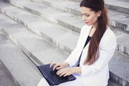 Beautiful brunette business woman in white suit with notebook on her lap, typing, working outdoors. Copy spaceの写真素材