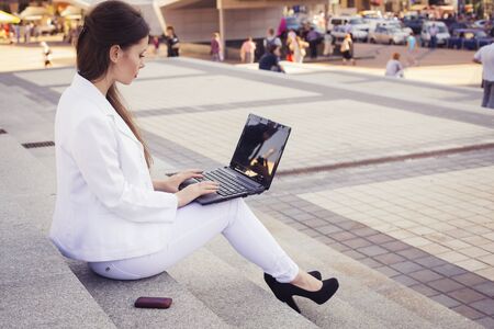 Beautiful brunette business woman in white suit with notebook on her lap, typing, working outdoors....