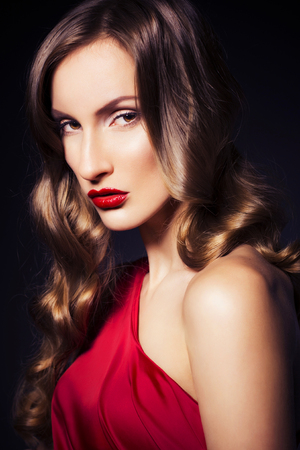 Brunette beautiful luxury woman in red dress with clear skin and evening dark make up: green cat eye and brown eyeshadows. Waved hairstyle. Dark backgroundの写真素材