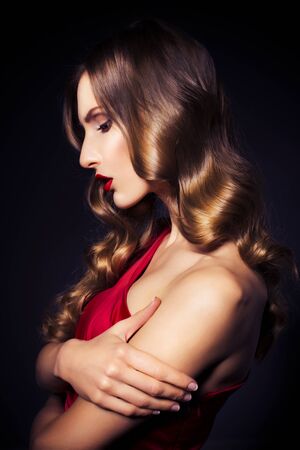 Brunette beautiful luxury woman in red dress with clear skin and evening dark make up: green cat eye and brown eyeshadows. Waved hairstyle. Dark backgroundの写真素材